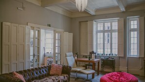 Una habitación con un sofá de cuero, un sillón de peluche rojo, una mesa de madera y una silla blanca. Se observan varias puertas y ventanas con contraventanas.