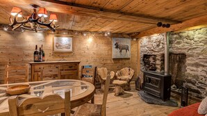 A rustic wooden interior with a round dining table, wooden chairs, a fireplace, and a stone wall. - Chalet Pele - Alpes Agency - Central Chamonix - Sleeps 11 (Chamonix)