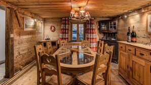 A rustic wooden interior with a round dining table surrounded by wooden chairs, a chandelier, and a kitchen area with wooden cabinets and a wine bottle. - Chalet Pele - Alpes Agency - Central Chamonix - Sleeps 11 (Chamonix)