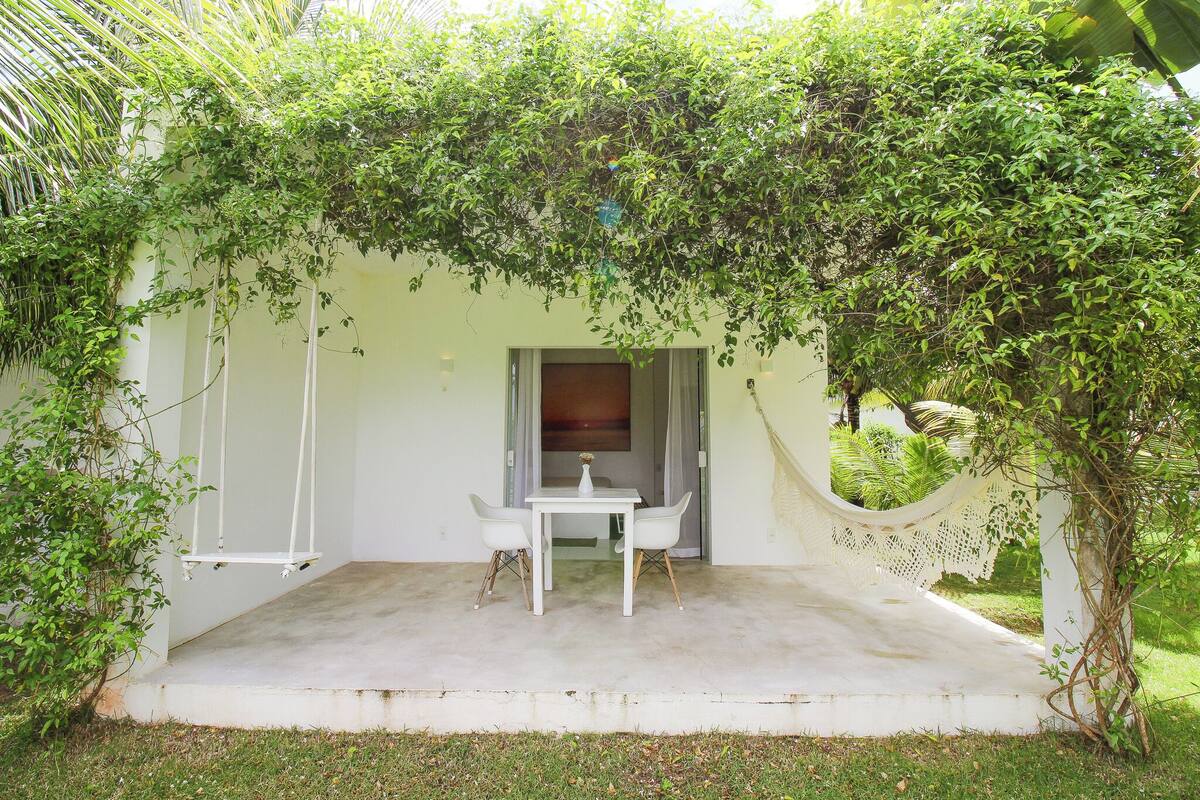 Un espacio al aire libre techado con una mesa y sillas, rodeado de exuberante vegetación y una hamaca.
