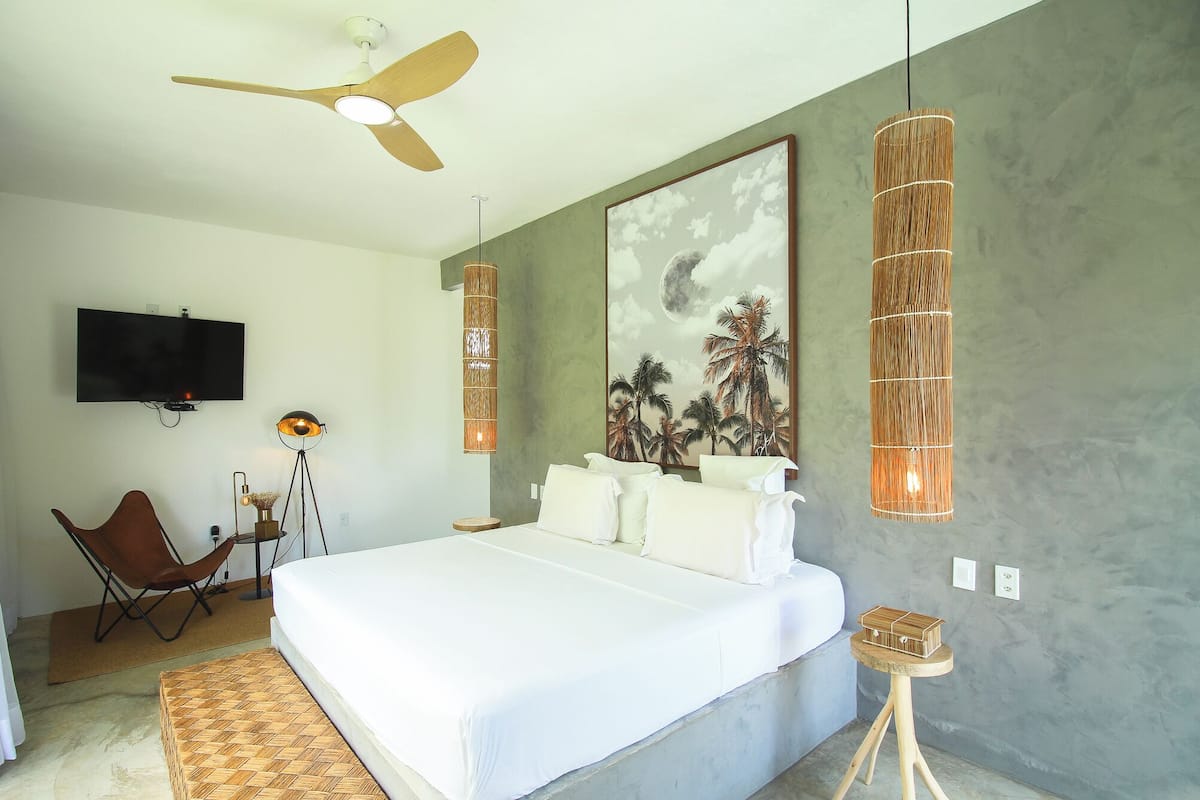 Un dormitorio moderno con una cama grande, ventilador de techo, televisor montado y un cuadro con palmeras y una luna.