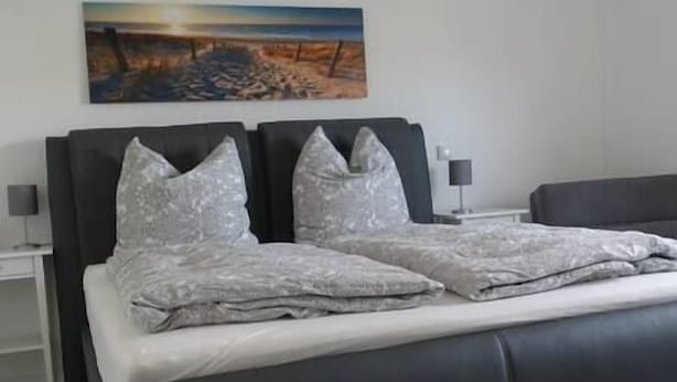Una cama doble con una colcha y almohadas estampadas, una mesita de noche con una lámpara y un cuadro de una playa en la pared.