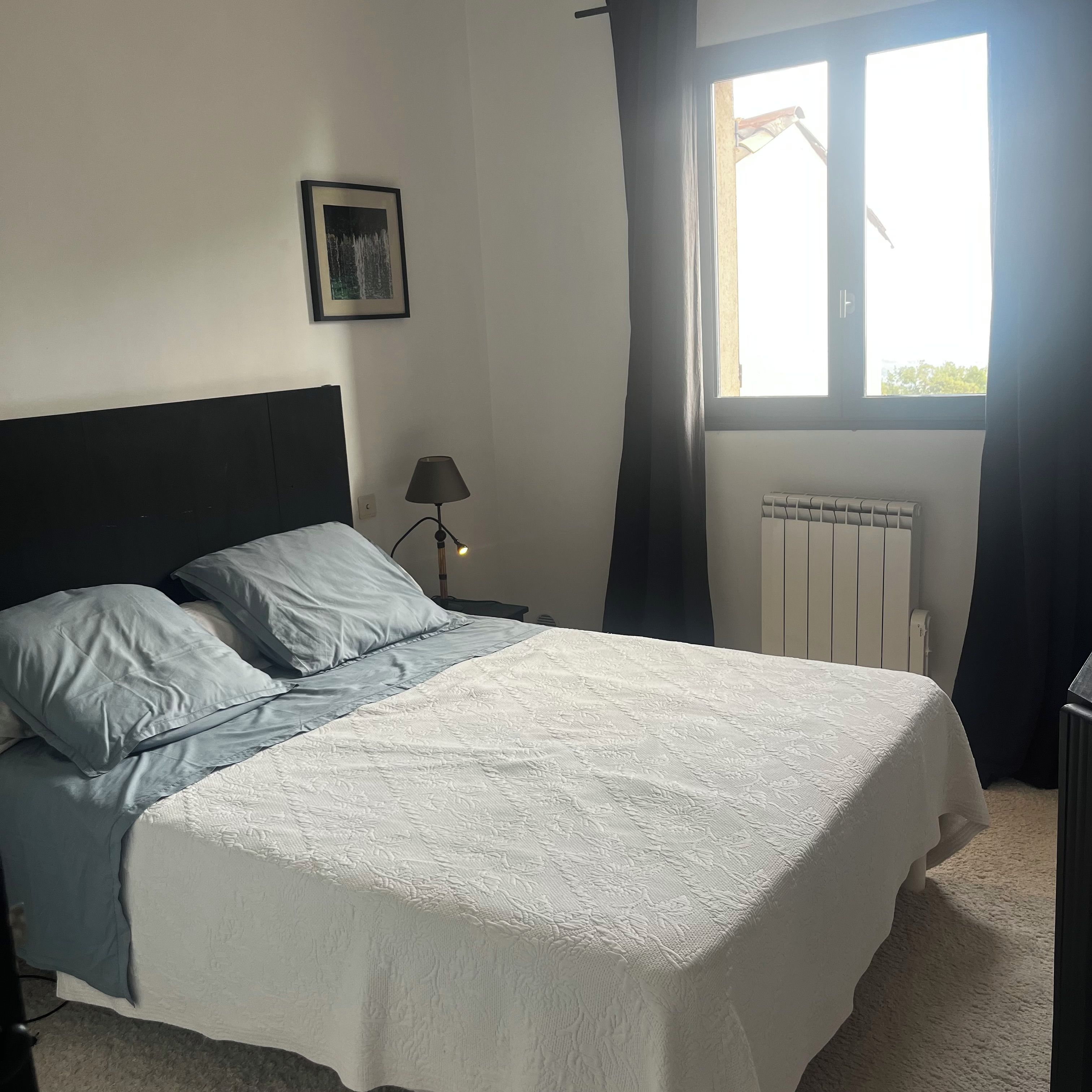 A bedroom with a bed, a nightstand, a lamp, a window, and a radiator.