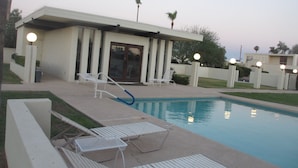 Un espace piscine comprenant des chaises longues, une piscine et un bâtiment doté d’une porte et de fenêtres.