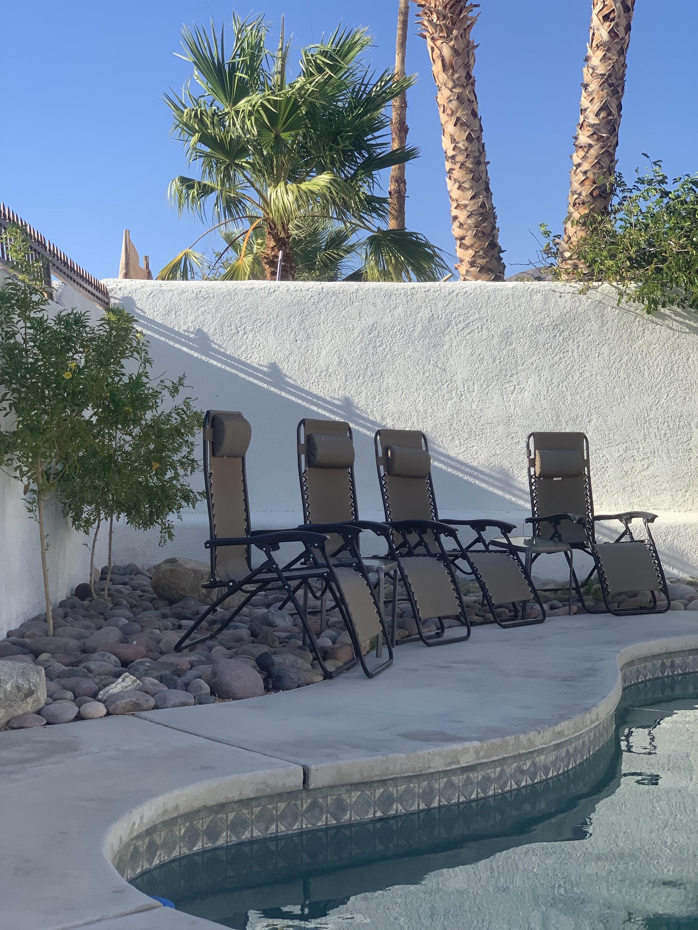 Un área junto a la piscina con sillones reclinables, una pared blanca y palmeras.