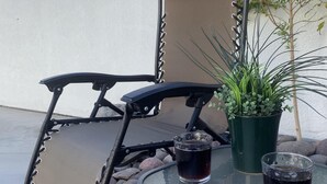 A patio chair with a cushion, a glass table, two glasses of dark liquid, a potted plant, and a small tree.