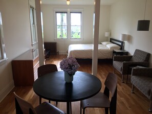 A room with a round table, two chairs, a bed, a nightstand, a lamp, and a window with a view of greenery. - Stunning Capitol Hill View Apartment (Seattle)