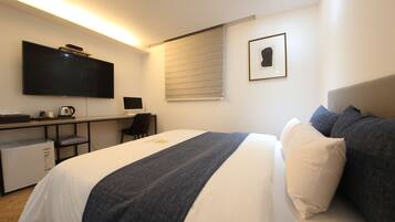 A hotel room with a bed, a desk with a computer, a television, and a window with blinds.