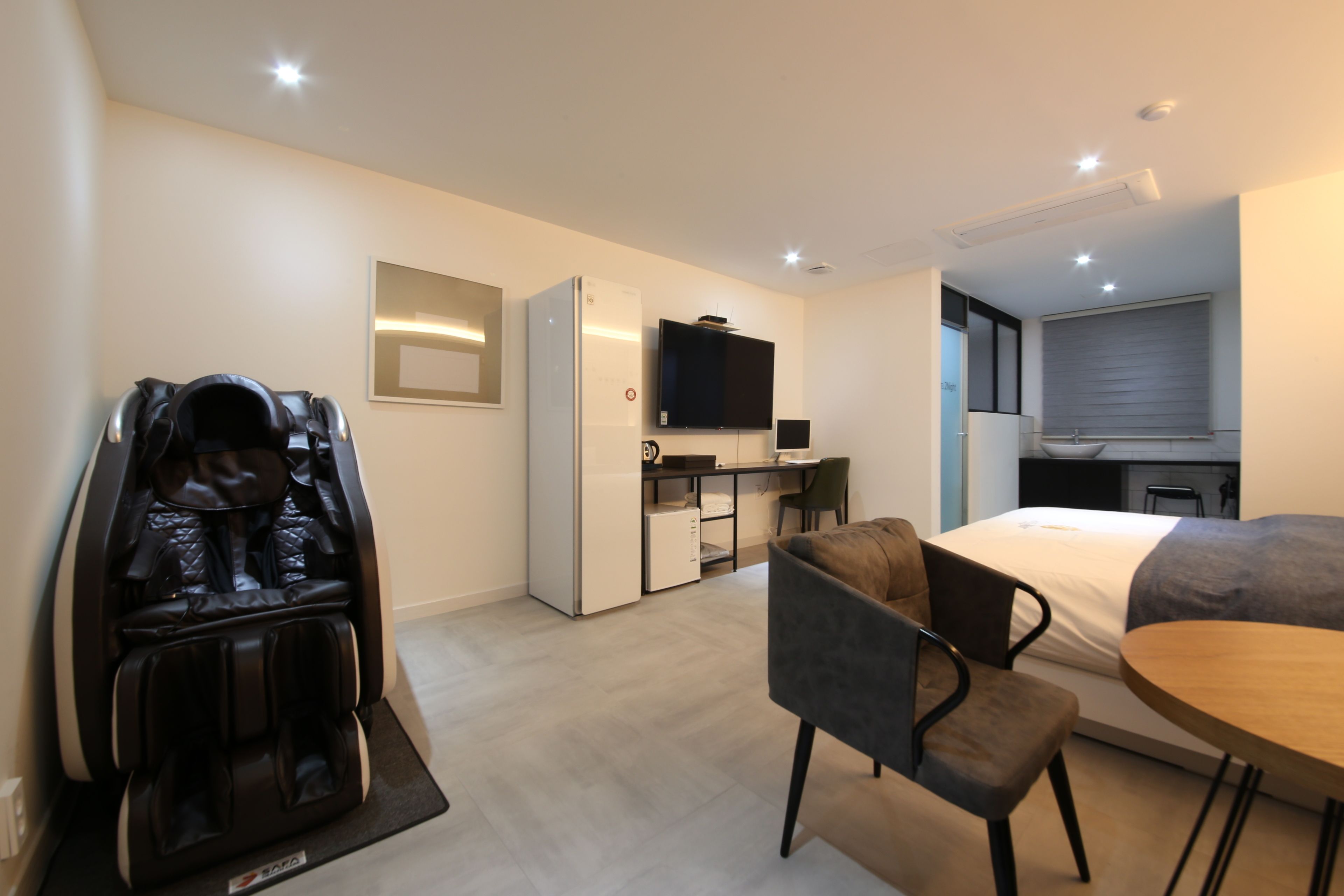 Una habitación de hotel moderna con sillón de masaje, cama, mesa de comedor, pequeña cocina y televisor montado en la pared.