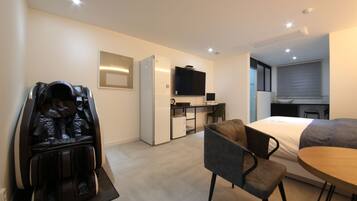 Una habitación de hotel moderna con sillón de masaje, cama, mesa de comedor, pequeña cocina y televisor montado en la pared.