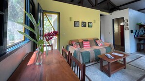 A cozy living room with a wooden table, a sofa with cushions, a large window with a view of greenery, and a ceiling with exposed beams.