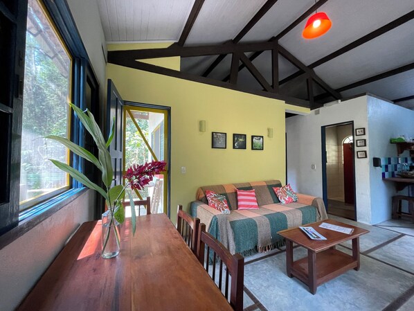 Uma sala de estar acolhedora, com uma mesa de madeira, um sofá com almofadas, uma janela grande com vista para o verde e um teto com vigas aparentes.