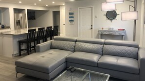 Ein modernes Wohnzimmer mit einem Ecksofa, einem Essbereich mit Stühlen, einer Küche mit Kühlschrank und einem Deckenventilator.