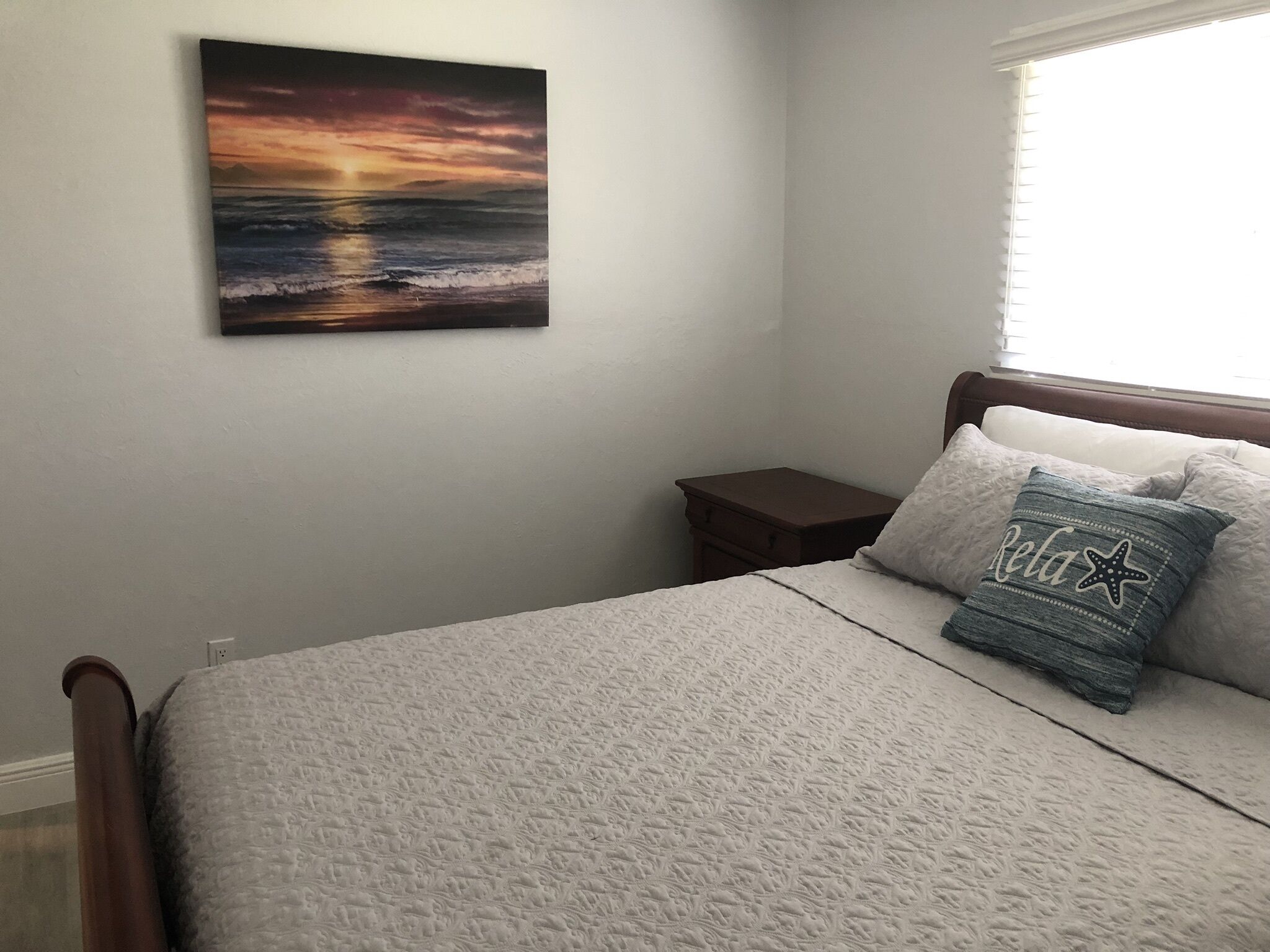 Un dormitorio con cama, mesita de noche, ventana y un cuadro de atardecer en la pared.