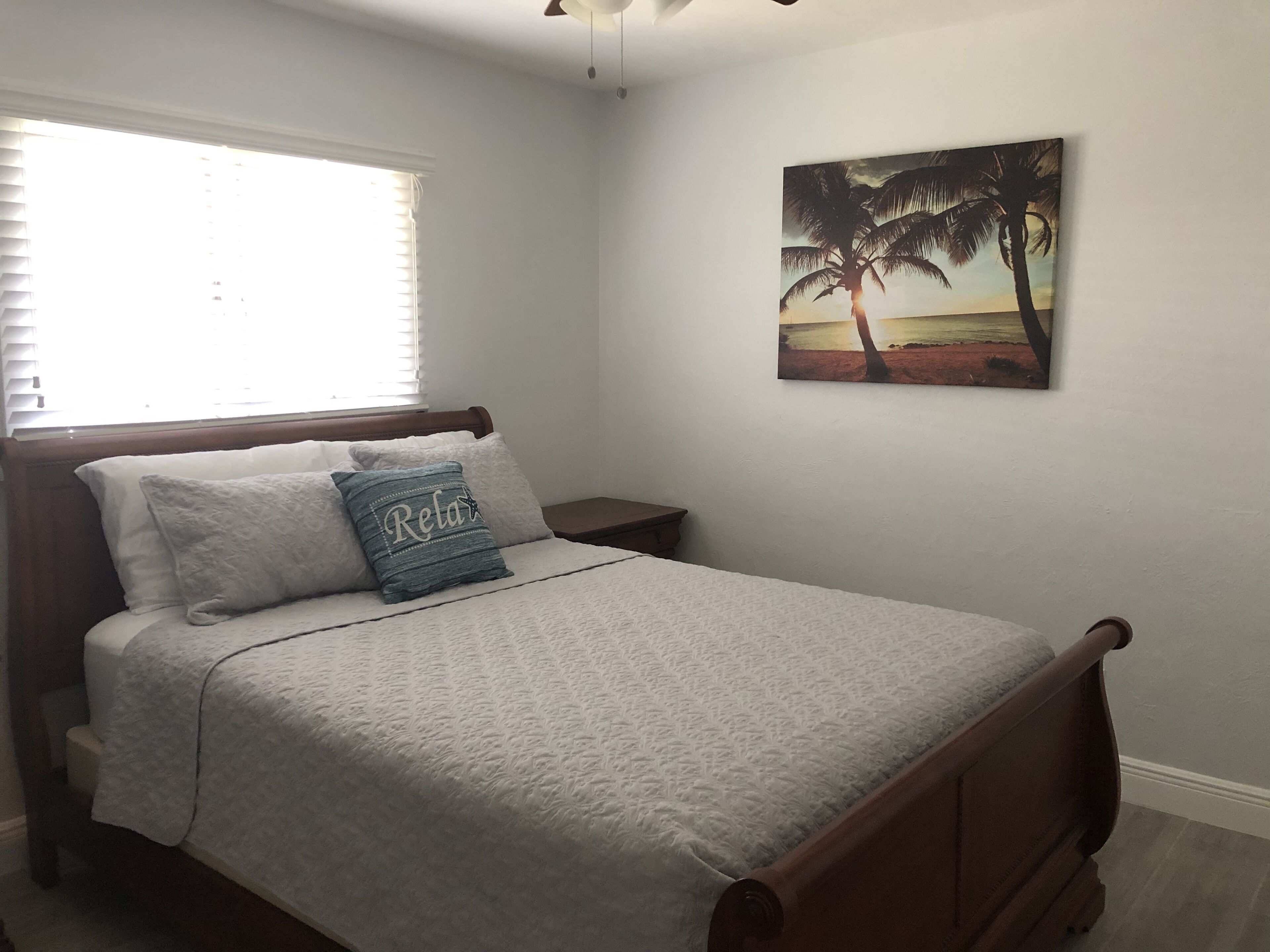 Une chambre à coucher comprenant un lit, une table de chevet, une fenêtre avec des stores et un tableau représentant un palmier accroché au mur.