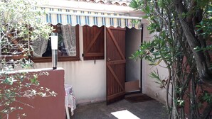 A narrow outdoor corridor with a wooden door, a small table with a checkered tablecloth, and a window with patterned curtains.