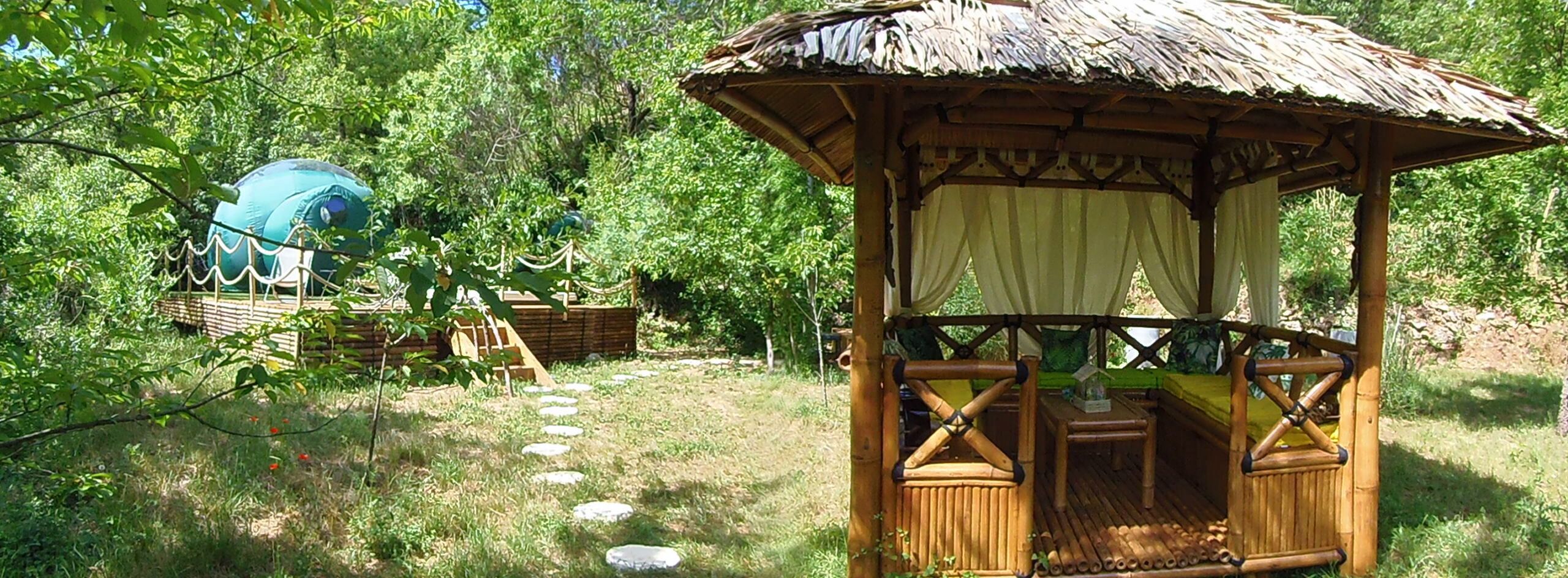 A rustic wooden gazebo with a thatched roof, surrounded by a natural setting with trees and a clear sky.
