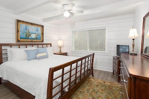 A bedroom with a wooden bed, a ceiling fan, a window with blinds, a bedside table with a lamp, and a dresser.