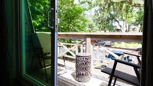 Una terraza de madera con una mesa y sillas de metal, un cubo de basura metálico y vistas a coches aparcados y árboles.