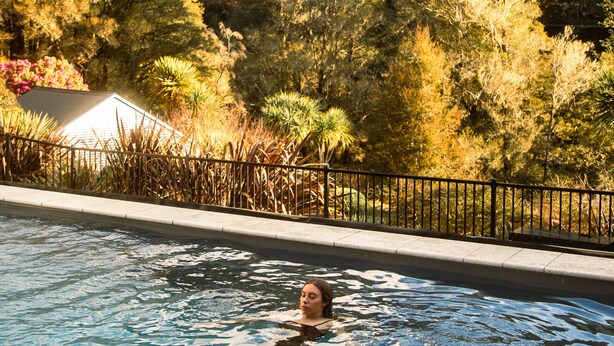 A swimming pool with a person in the water, surrounded by trees and a mountain in the background. - Maruia River Retreat (Shenandoah)