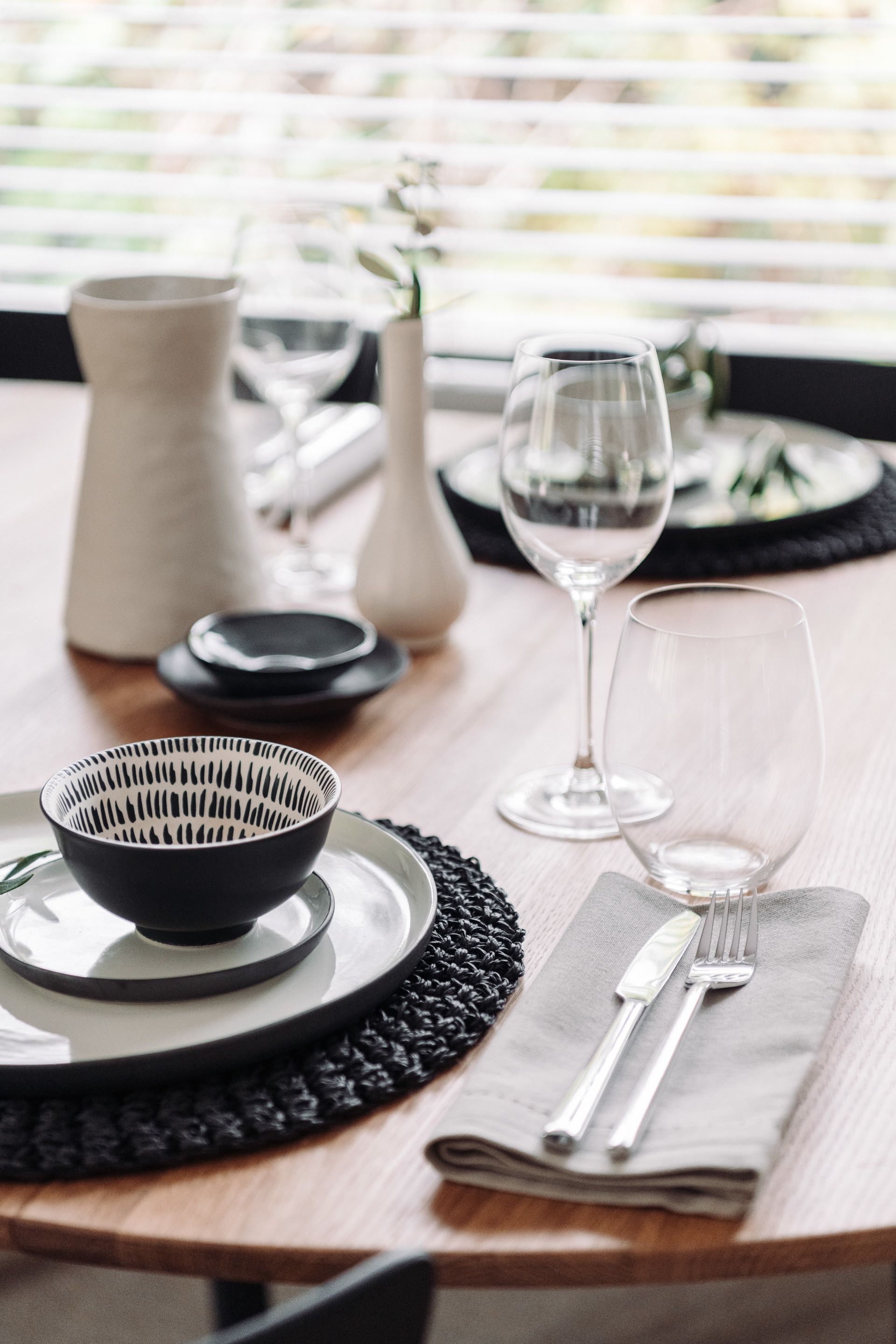 Ett matbord dukat med en svart skål, två vinglas, en kniv och en gaffel på en servett samt en vas med blommor.