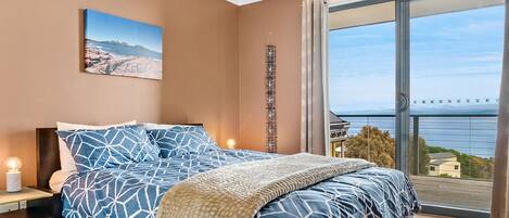 A bedroom with a large bed, a nightstand, a wall-mounted clock, and a window with a view of the sea.