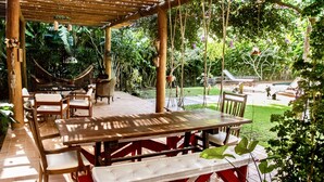 An outdoor patio with wooden tables and chairs, surrounded by lush greenery and a hammock. - Beautiful house in Ilhabela (Ilhabela)