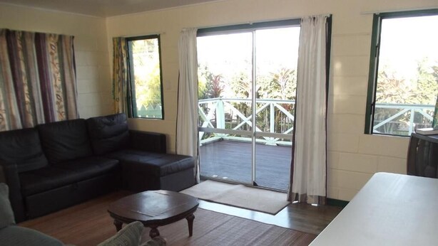 A living room with a sofa, armchair, and a coffee table. There is a sliding glass door leading to a balcony with a railing. - Green Lodge Holiday Homes (Nuku'alofa)