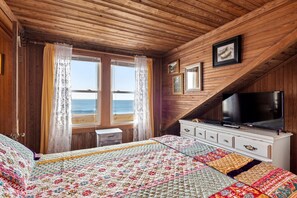 A cozy bedroom with a wooden ceiling, a large bed with a colorful quilt, a TV on a dresser, and a view of the ocean through a window.