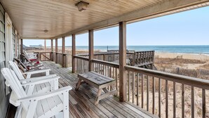 Terraza de madera con sillas Adirondack blancas y una mesa de picnic, con vistas a la playa.