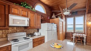 Una cocina con armarios de madera, una estufa blanca, un microondas y un refrigerador. Se ve una ventana con vista a la playa.