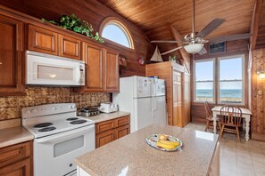 A kitchen with wooden cabinets, a white stove, a microwave, and a refrigerator. There is a window with a view of the beach.