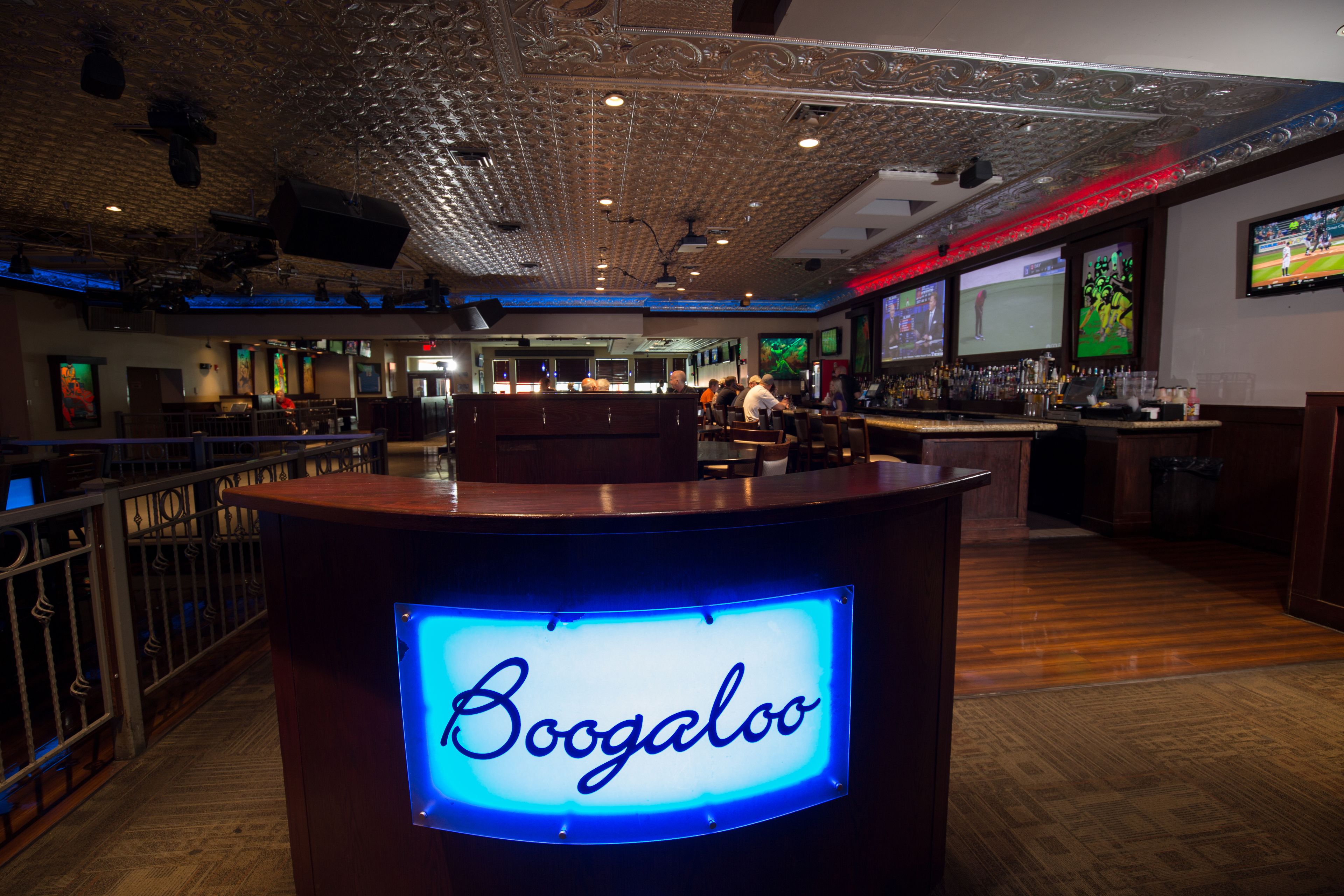 A bar area with a neon sign reading "Boogaloo," wooden flooring, and multiple screens displaying sports.