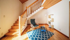 Una escalera de madera lleva a un dormitorio en un altillo con un tapete azul estampado, una silla y una mesa de fútbol.