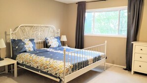 Un dormitorio con estructura de cama metálica, una ventana con cortinas, una mesita de noche y un buró.