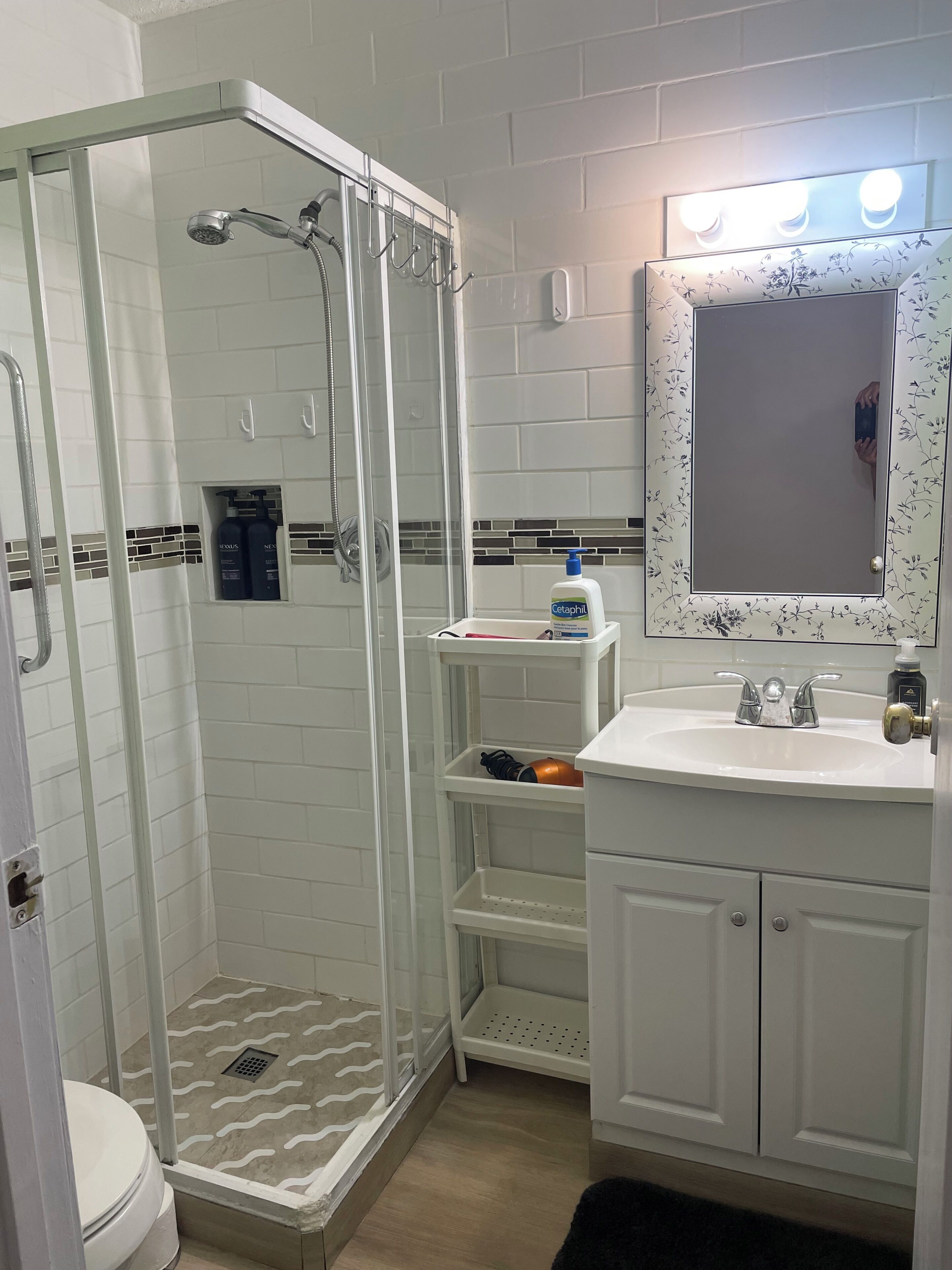 A bathroom with a glass-enclosed shower, a toilet, a sink with a mirror, and a shelf with toiletries.