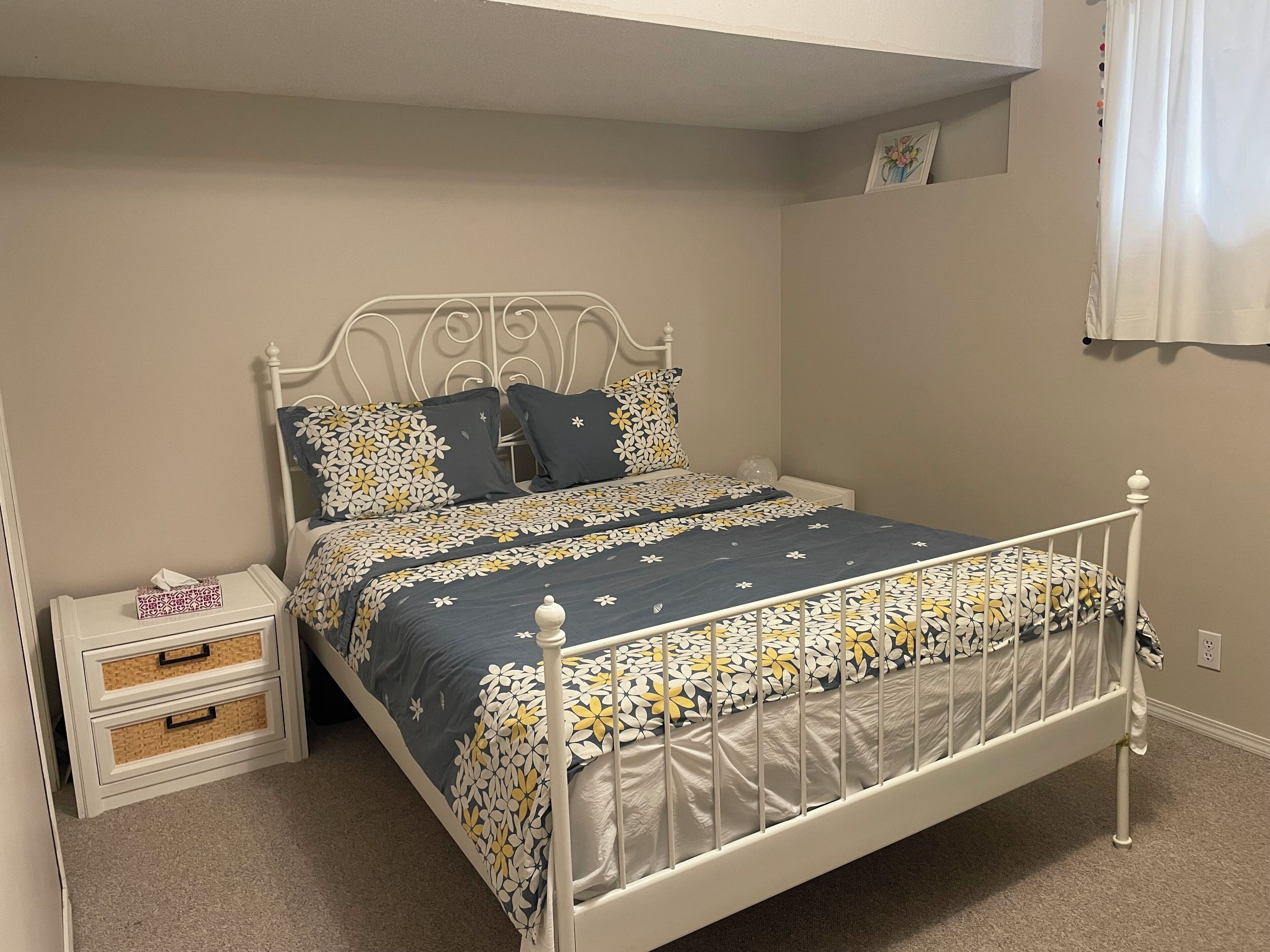 A bed with a floral bedspread, a nightstand with drawers, a wall-mounted picture, and a window with curtains.