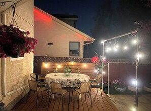 An outdoor patio with a round table and chairs, string lights, and a wooden bench.