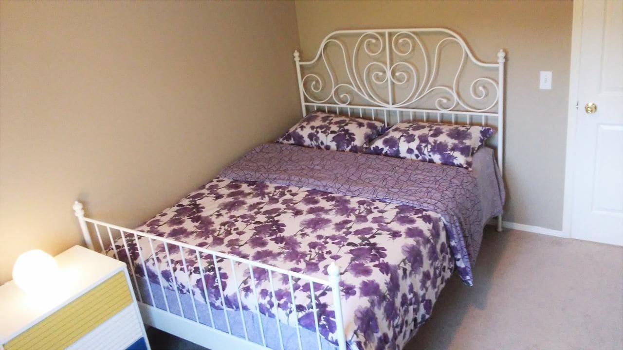 A single bed with a metal frame and floral bedding, a bedside table with a lamp, and a door to the right.