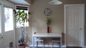 A room with a white door, a window with blinds, a wall clock, a potted plant, and a small table with two chairs.