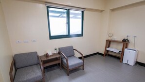 Una habitación pequeña y bien iluminada con dos sillones grises tapizados, una mesita de madera, un estante con un electrodoméstico blanco y una ventana con persianas.
