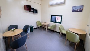 Una habitación pequeña y bien iluminada, con cuatro mesas y sillas, un equipo de aire acondicionado, una ventana con vistas y un estante con un jarrón.