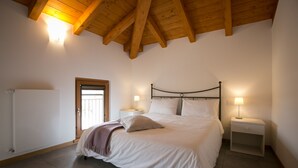 Een slaapkamer met een houten plafond, een bed met wit beddengoed, twee nachtkastjes met lampen en een deur links.
