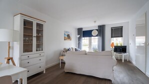 Uma sala de estar com sofá, aparador com portas de vidro, luminária, televisão e janela com cortinas.