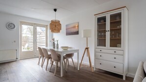 Uma sala de jantar com uma mesa branca, cadeiras e um abajur. Há um armário com portas de vidro e um relógio na parede.