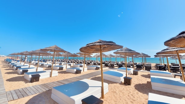 Une plage bordée de rangées de chaises longues et d’ombrelles en chaume, une terrasse en bois et un ciel bleu azur.