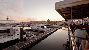 Un port de plaisance avec des bateaux amarrés, une zone abritée où se trouvent des personnes, et une étendue d’eau calme qui reflète le ciel.