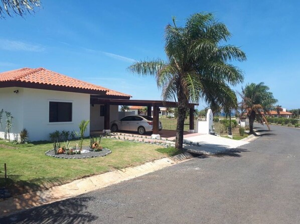 A single-story house with a red-tiled roof, a covered carport, and a well-maintained lawn with small plants and a palm tree. - Casa Boroda - Andromeda Pedasi - In Park with Swimming pool and Beach (Pedasi)
