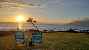 Zwei gestreifte Liegestühle blicken auf einen Sonnenuntergang über dem Meer, mit einem Baum und einer felsigen Küstenlinie.