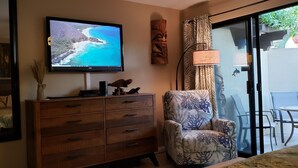 Ein Zimmer mit Fernseher, der eine Strand Szene zeigt, einem Holzschrank, einem gemusterten Sessel und einer Glas-Schiebetür, die nach draußen führt.